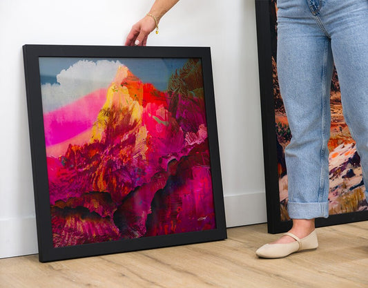 Square acrylic photo print of an abstract mountain scene leaning against a wall beside a woman’s legs.