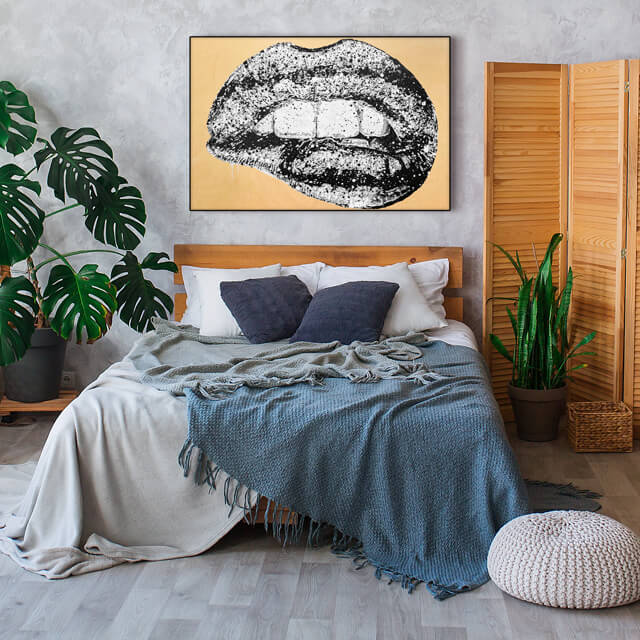 Black and white painting of lips on yellow background hangs above bed in boho bedroom.