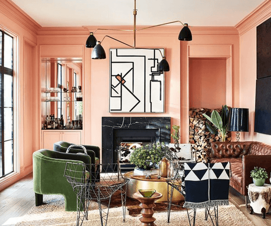Colorful living room with pink walls, green velvet chairs, a brown leather sofa, and a black and white geometric artwork above a black fireplace.