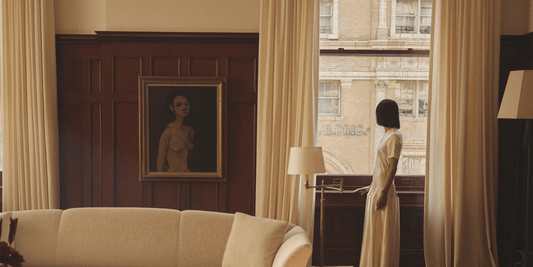 woman in white dress looking out window of art deco style apartment with portrait on the wall and white couch in foreground