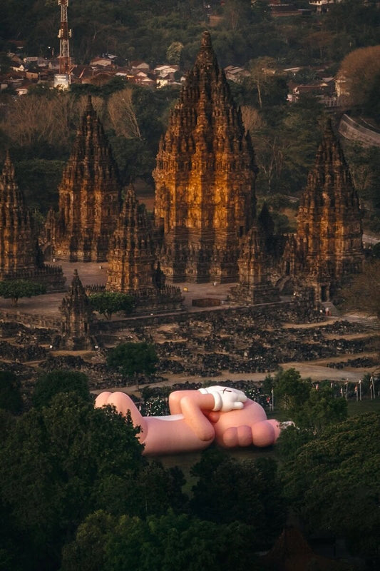 KAWS:HOLIDAY scuplture laying in the grassy lawn beside 9th century Indonesian temple, Prambanan Temple