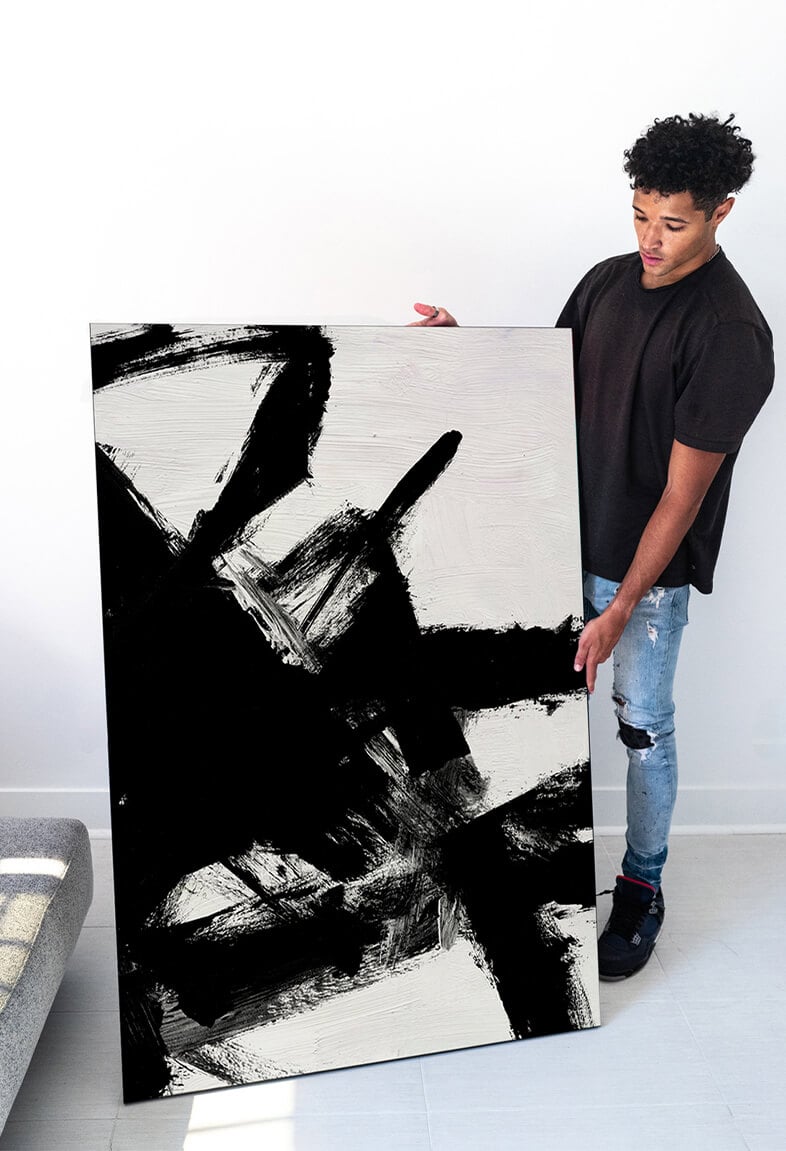 Huge black and white abstract painting of white background layered with black thick brushstrokes is held by Black man in clean white living room.