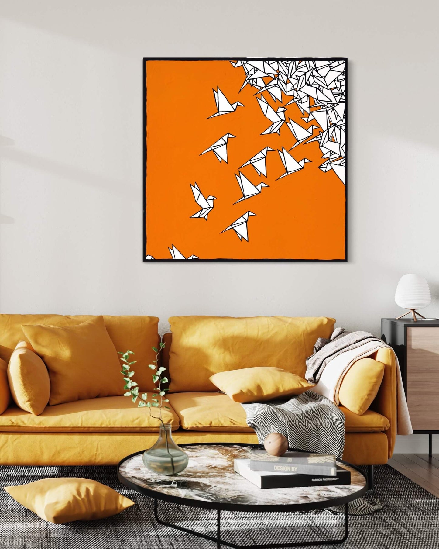 Boho modern art hung on a light grey wall above a large mustard colored couch. The boho art features a bright orange background with origami birds flying across