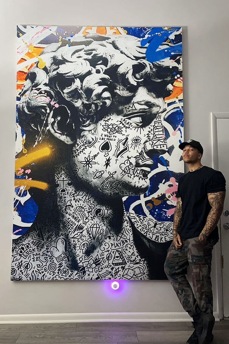 Oversized pop art of Michelangelo's David featuring blue graffiti spray paint surrounding the doodle covered face hangs in living room next to man with tattoo sleeves.