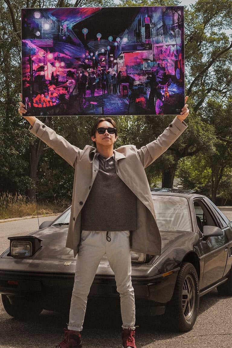 Man stands in front of car and holds large magenta and navy wall art overhead featuring abstract drawing of city street at night.