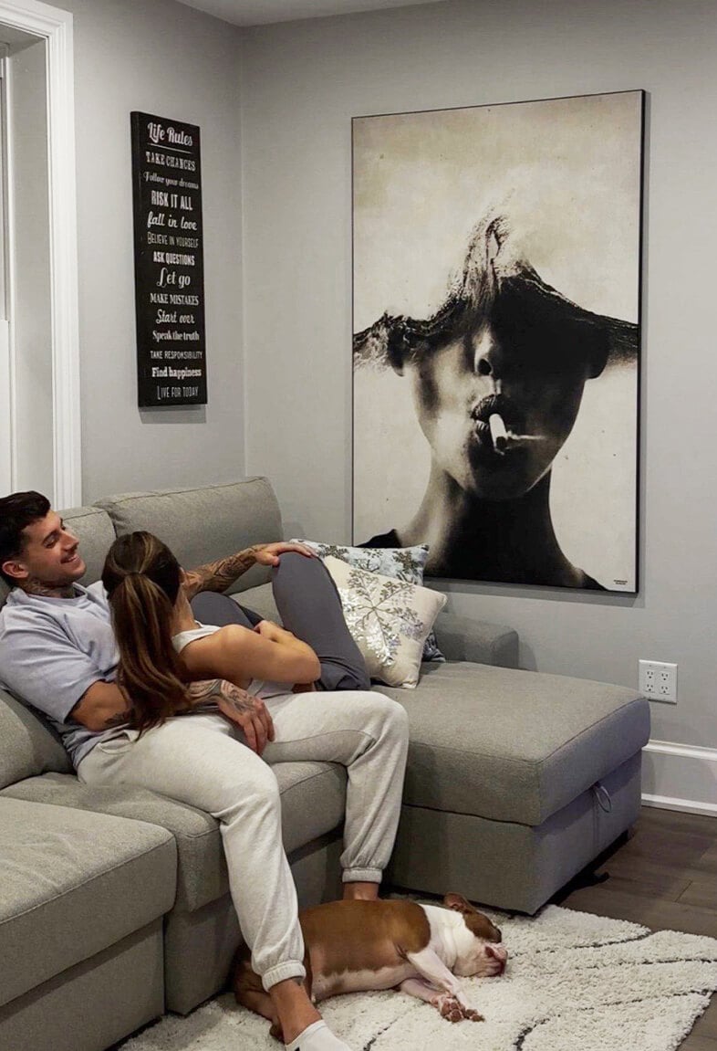 Huge minimal black and white photo of person smoking with waves for the top half of their head hangs on neutral wall in living room with large grey section where couple sits and cuddles.