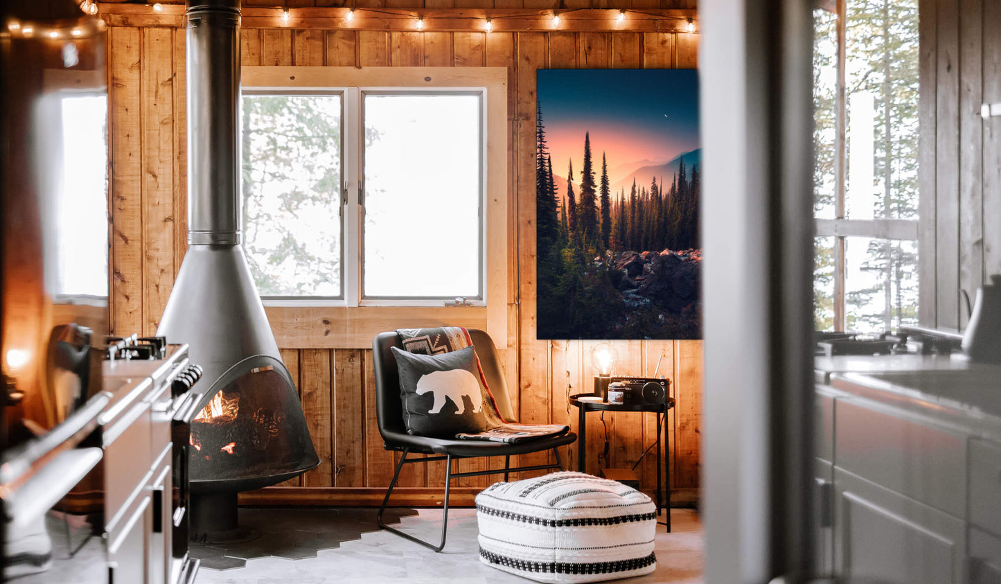 Oversized evergreen forest vertical wall art of sunset on the mountains hangs on wood paneled wall in rustic cabin.