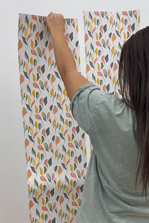 Person hanging a sheet of colorful leaf-patterned wallpaper on a white wall. loading=