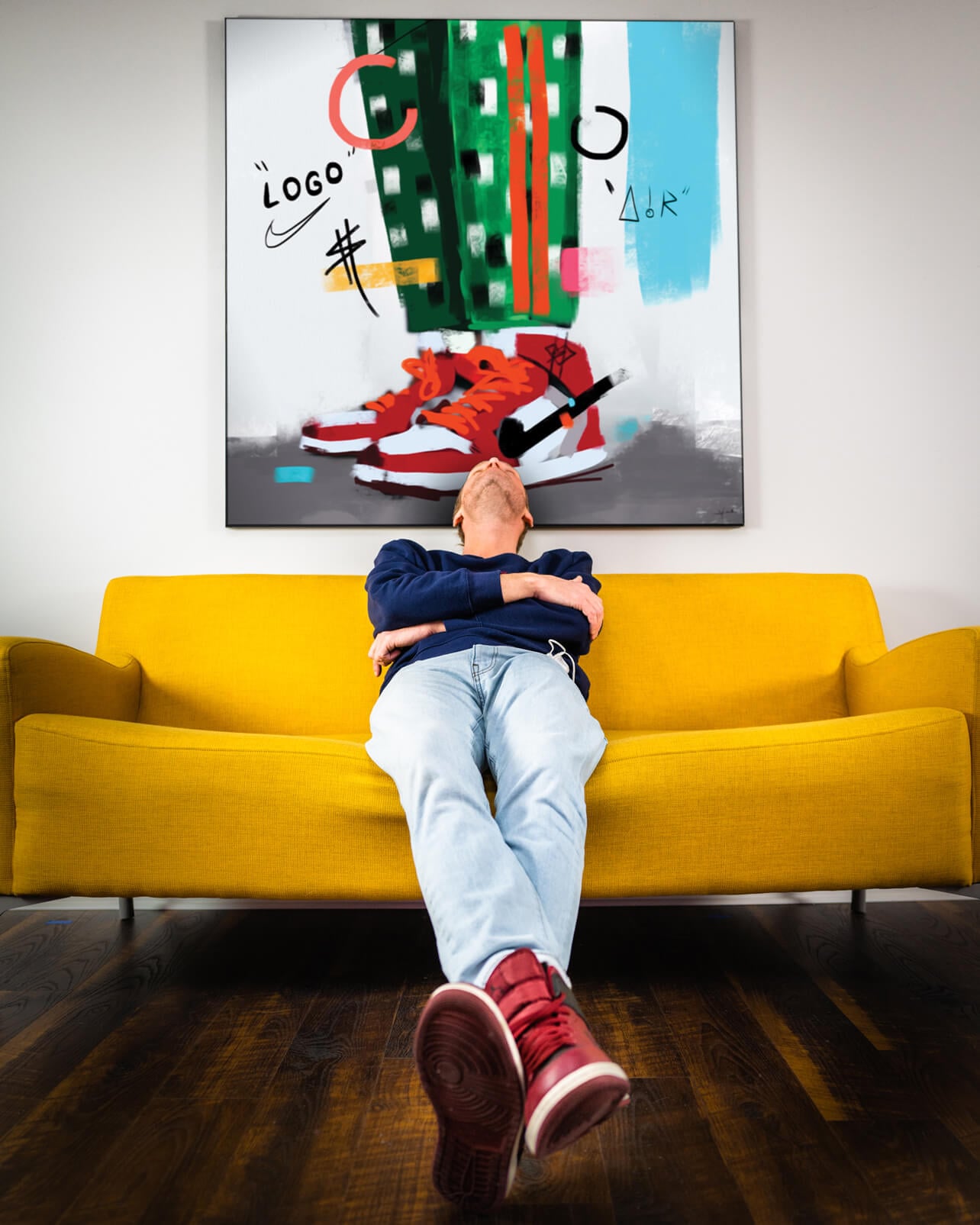 Huge colorful hip hop art of digitally drawn Air Jordans on a person wearing green and white checkered pants hangs above yellow couch. On the couch, a man casually leans back, chin tilted towards the sky. On his feet are red Air Jordans, which are similar to the sneakers drawn in the wall art.