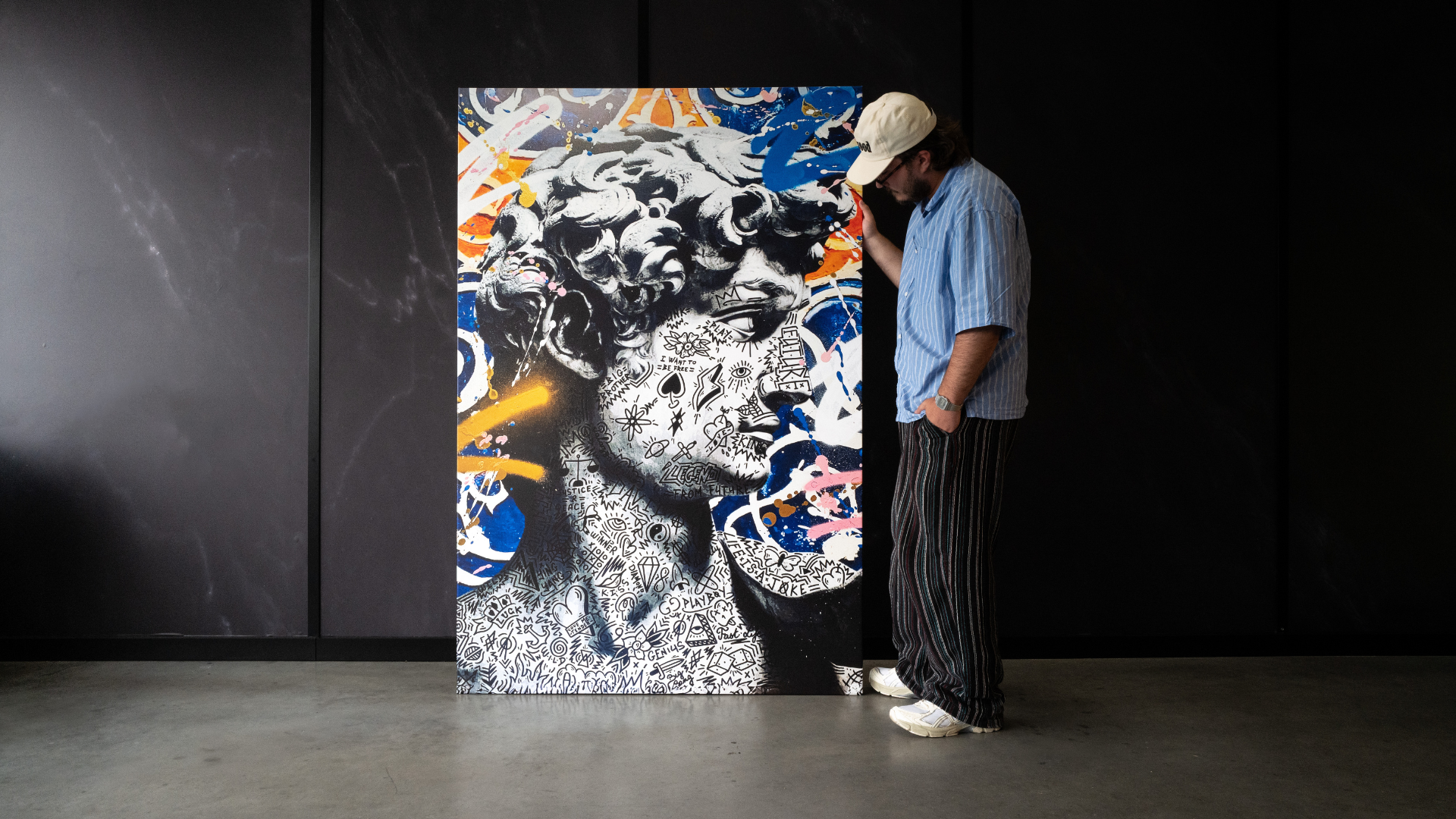 A man in a light blue shirt, white cap, and striped pants stands beside a large, highly detailed black-and-white portrait painting with vibrant blue, orange, and pink graffiti-style accents against a dark gallery wall.
