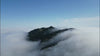 A wide shot looking out across a dense sea of low-lying white clouds, with a dark, forested mountain peak breaking through the fog under a clear blue sky.