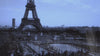 A vintage-styled, cool blue-toned shot of the Eiffel Tower rising above a crowded plaza in Paris, with people gathered around a large fountain in the foreground under an overcast sky.