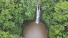 A high-angle aerial view of a narrow waterfall cascading down a rocky cliff into a brown pond, surrounded by a dense green tropical forest.