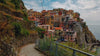 A wide shot of a colorful coastal town built onto a steep cliff side overlooking a blue sea, with a paved walking path and metal railing in the foreground under a cloudy sky.