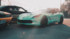 A low-angle, front-facing shot of a mint-green Corvette parked on an asphalt lot next to an orange Dodge Challenger and a black sedan, with warm sunlight flaring from the upper right.