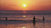 A wide, low-angle shot of three surfers sitting on their boards in the ocean waves, silhouetted against a brilliant, glowing orange sky as the sun rises over the horizon.