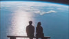 The silhouette of a man and a woman sitting side-by-side on a bench, looking out at a large view of planet Earth with a bright sun reflection glittering across the ocean surface.