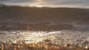 A close-up, low-angle shot of gentle water waves lapping onto a pebbled shoreline, with the setting sun reflecting a bright, golden shimmer across the surface of the water.