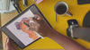 An overhead shot of an artist drawing a detailed portrait on a digital tablet with a stylus, set against a bright yellow desk with a coffee mug and accessories.
