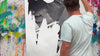 A close-up shot of an artist creating a mixed-media canvas collage with ripped posters, spray paint, and a printed rose pattern in shades of pink, white, and red.