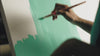 A close-up, high-angle shot focusing on an artist's hand using a paintbrush to fill in a large white canvas with strokes of a solid, bright teal paint.