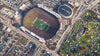 A high-angle, birds-eye aerial view of a packed football stadium during a game, with a passenger airplane flying directly over the field and crowded parking lots surrounding the arena.