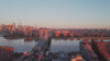 A wide aerial view of the Williamsburg Bridge crossing a wide river into a hazy, warm-toned city skyline at sunset, with golden light reflecting off the skyscrapers.