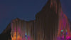 A digital glitch art effect applied to the silhouette of a mountain range at dusk, with vertical streaks of pink, orange, and blue light dripping down from the dark peaks.