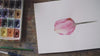 A top-down view of a completed pink watercolor tulip painting on white paper, resting on a messy wooden desk next to a well-used paint palette and brushes.