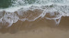 A top-down, aerial view of deep turquoise ocean waves crashing onto a tan sandy beach, creating white seafoam across the shoreline.