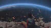 A back-view shot of a person sitting on a rocky, flower-lined ledge, looking out over the curved horizon of planet Earth from space, with city lights visible on the dark side of the globe.
