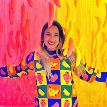 Female artist Jess Stempel poses in front of a pink and yellow backdrop with corresponding colored bananas hanging from the ceiling around her. She is wearing a colorful sweater and grins widely in this playful and creative setting.