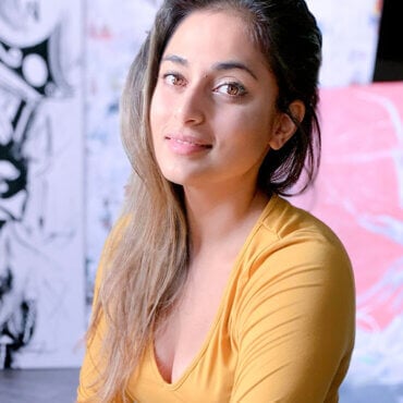 Female artist P Muir sits in her studio in front of a few of her paintings, wearing a yellow long sleeve shirt, and poses for Women's History Month.