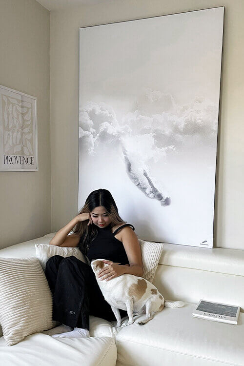 Oversized minimalist wall art of person sky diving through white clouds hangs above white couch in white living room. A woman dressed in black sits on the couch beneath the white minimalist wall art and pets her dog.