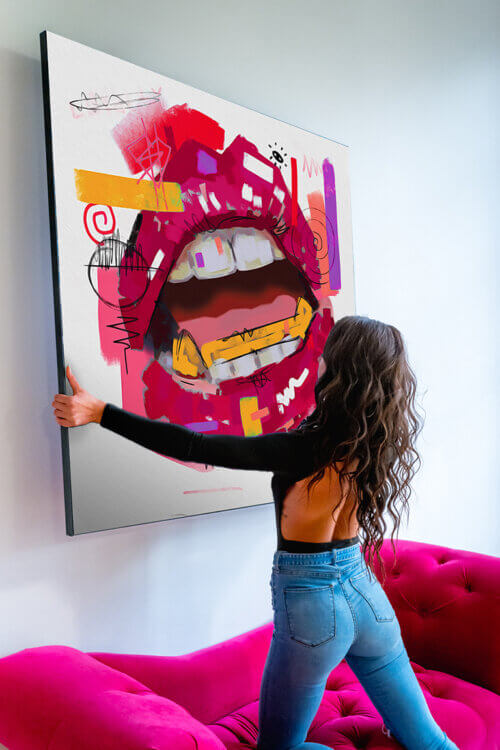 Woman kneels on magenta pink velvet couch and hangs large square wall art featuring magenta pink lips drawn in pop style.