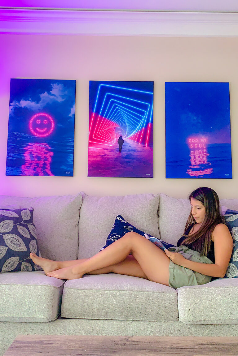 Set of three magenta and dark blue artworks featuring neon signs hang above neutral couch in modern woman's living room.