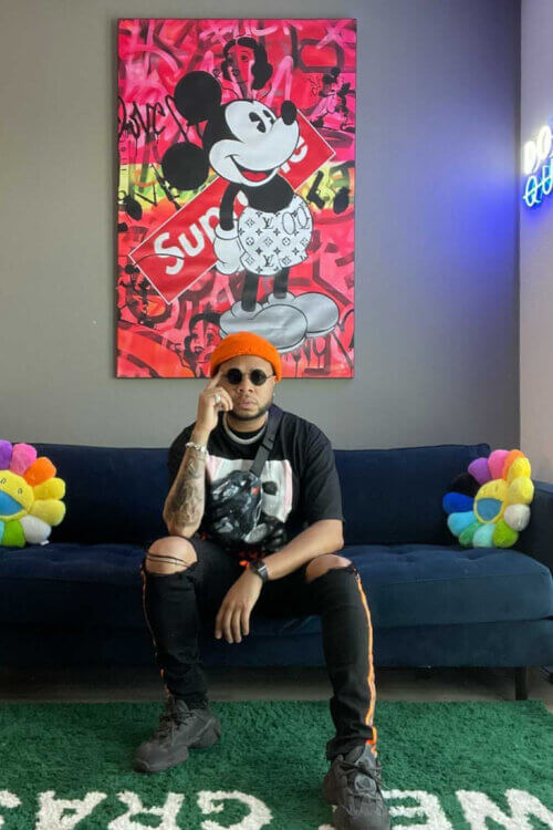 Cool man cave wall art with red graffiti background and Mickey Mouse cartoon wearing white designer Louis Vuitton shorts hangs above navy couch where man sits and poses for the camera.