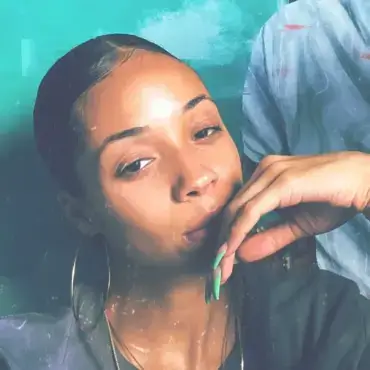 Black painter Kristen Campbell poses in front of a digital painting featuring a bright teal background and the back of a Black woman's head with curling rollers in her hair, wearing large gold hoop earrings and a sleek bun, for Women's History Month.