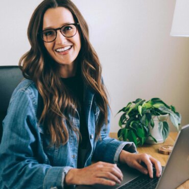 Female graphic designer Olivia Herrick sits in front of her laptop at her desk, wearing a dark blue jean jacket and glasses, while creating designs for Women's History Month