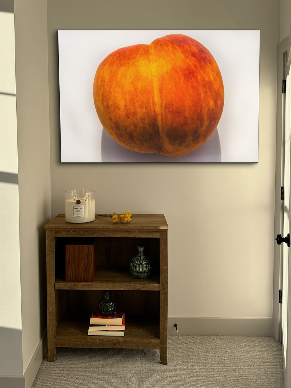 Large photo of a peach that mimics the shape of the peach emoji that is synonymous with a butt is shown on a white background and hangs on a neutral wall in a bedroom above shelf topped with Big Ass Candle in Sparkling Paloma scent.