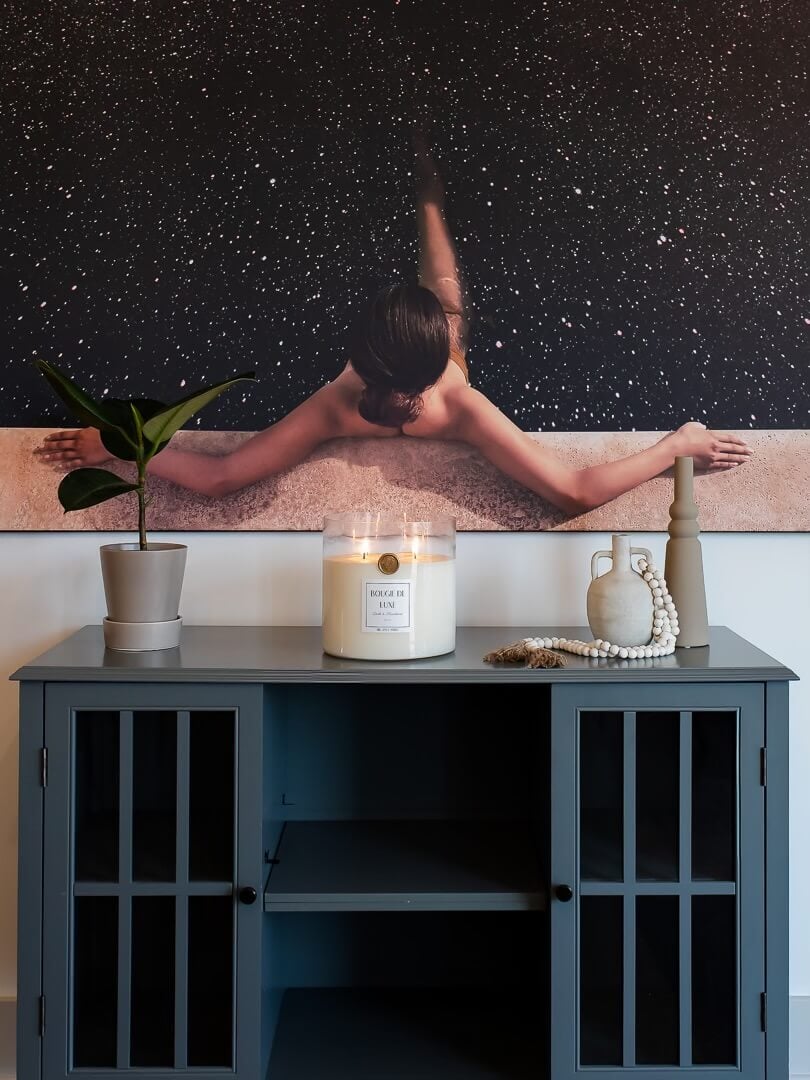 Big Ass Wall art from BIG Wall Décor featuring a naked woman hanging on the edge of a pool that is filled with stars and a dark sky instead of pool water hangs above cabinet in dining room that has a big ass candle sitting on top of it. The Big Ass Candle is in the scent Dark & Handsome. It is surrounded by other home decor, a small indoor rubber leaf tree plant and tow ceramic vases in light grey neutral colors.