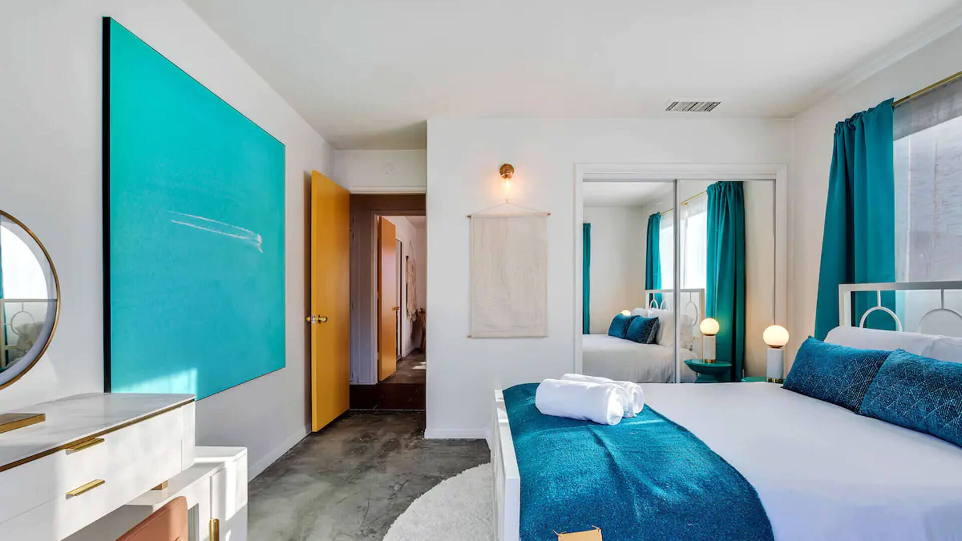 Large teal blue abstract art hangs on white wall at the foot of bed in neutral blue and white bedroom.