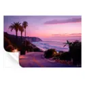 Coastal road at sunset with palm trees, rugged cliffs, and a pink and purple sky.