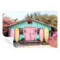 Turquoise beach hut with a pink door, surrounded by blooming flowers and colorful surfboards.