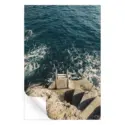 A coastal photograph featuring a stone staircase leading to a metal ladder descending into the deep blue sea with foamy waves.