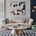 Modern minimalist maximalist living room with a cream tufted sofa, green velvet accent chair, geometric wall art, and a navy checkered rug