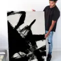 Huge black and white abstract painting of white background layered with black thick brushstrokes is held by Black man in clean white living room.