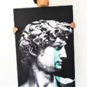 Oversized vertical black and white wall art of David of Michelangelo statue with pop of blue tears is held in white living room by Black man.