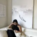 Oversized minimalist wall art of person sky diving through white clouds hangs above white couch in white living room. A woman dressed in black sits on the couch beneath the white minimalist wall art and pets her dog.