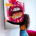 Woman kneels on magenta pink velvet couch and hangs large square wall art featuring magenta pink lips drawn in pop style.